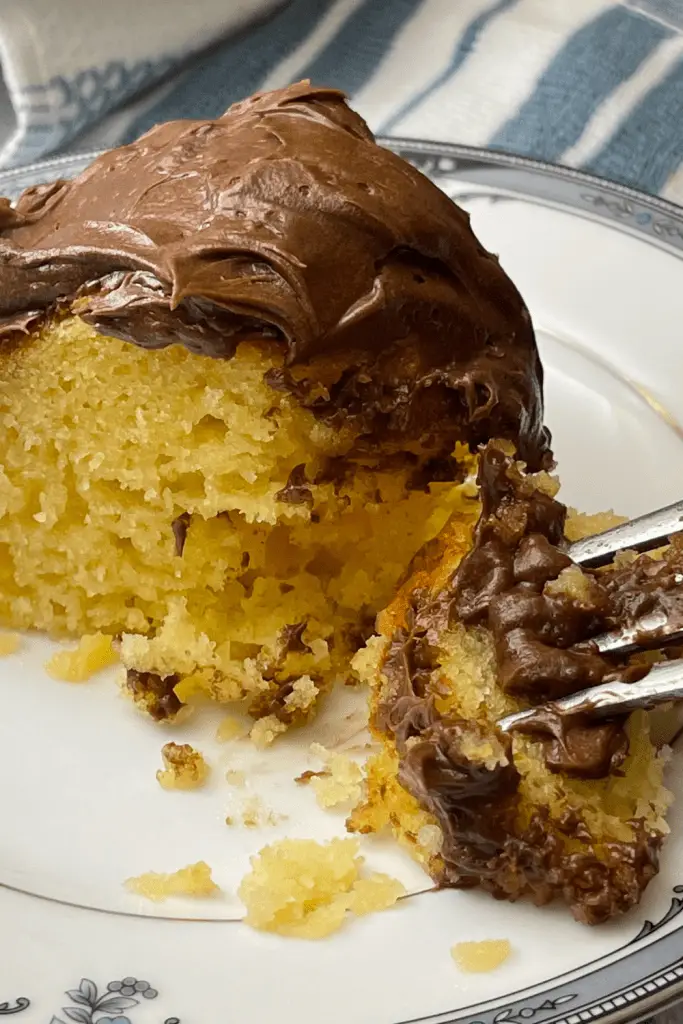 A piece of vanilla cake with chocolate buttercream frosting on a china plate with a bite of cake on a fork.